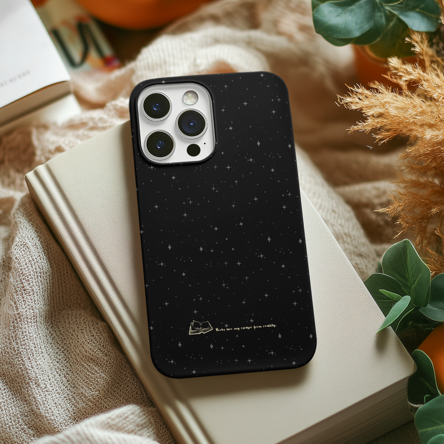 A black phone case with a starry night design, featuring scattered white stars and a gold illustration of an open book with the quote 'Books are my escape from reality.' The phone has multiple camera lenses at the top left corner.